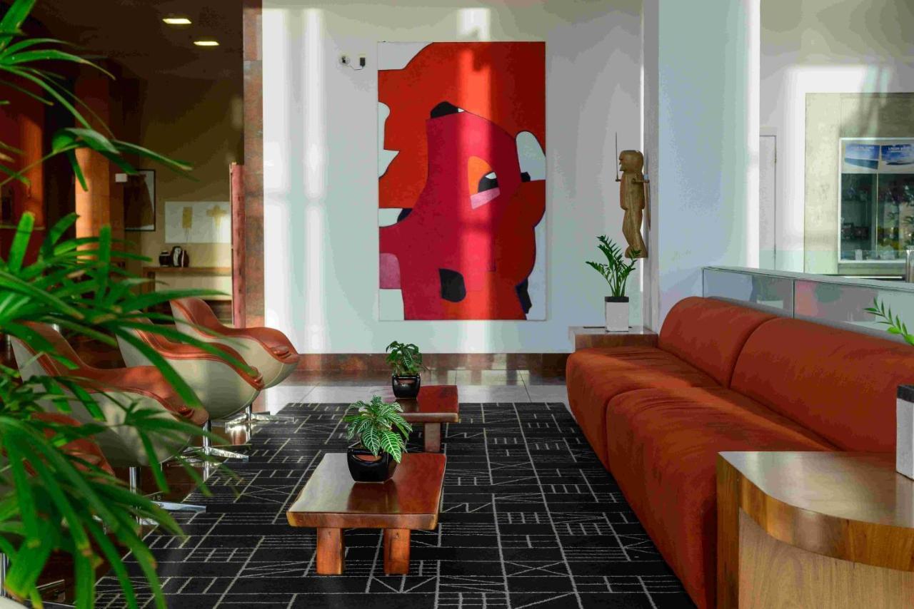 Hotel Luzeiros Sao Luis Eksteriør billede The photo depicts a modern interior space, likely a lounge or living area. In the foreground, there is a large orange sofa complemented by two small wooden coffee tables, each adorned with potted plants. The floor features a black and white patterned