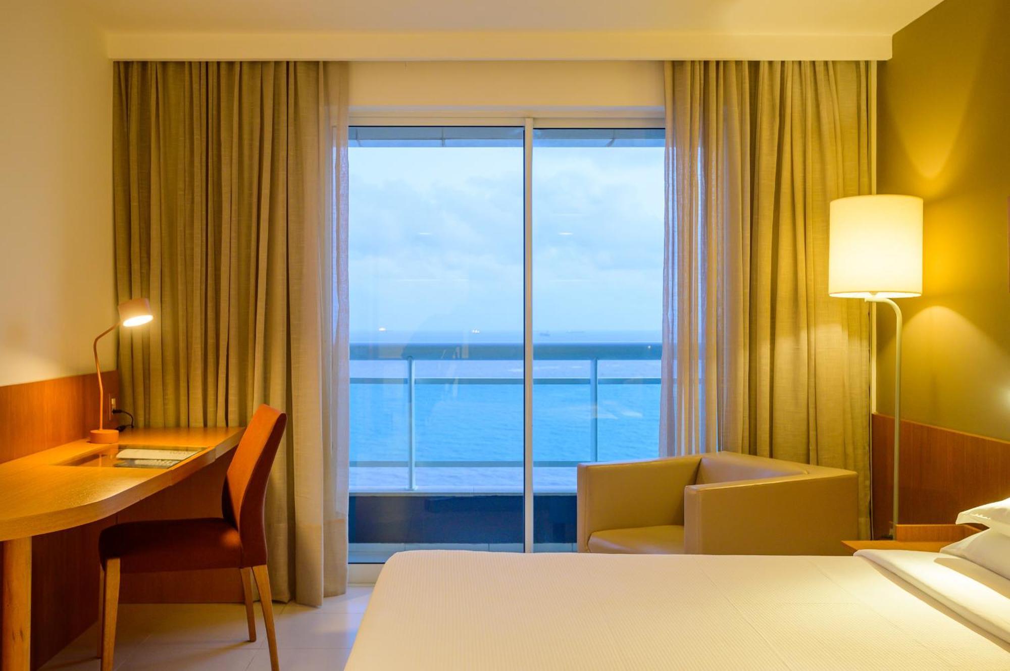 Hotel Luzeiros Sao Luis Eksteriør billede The photo shows a modern hotel room with a minimalist design. There's a large bed with white linens, and beside it, a small desk with a chair, illuminated by a lamp. A cozy armchair is positioned near a large window, which offers a view of the ocean 
